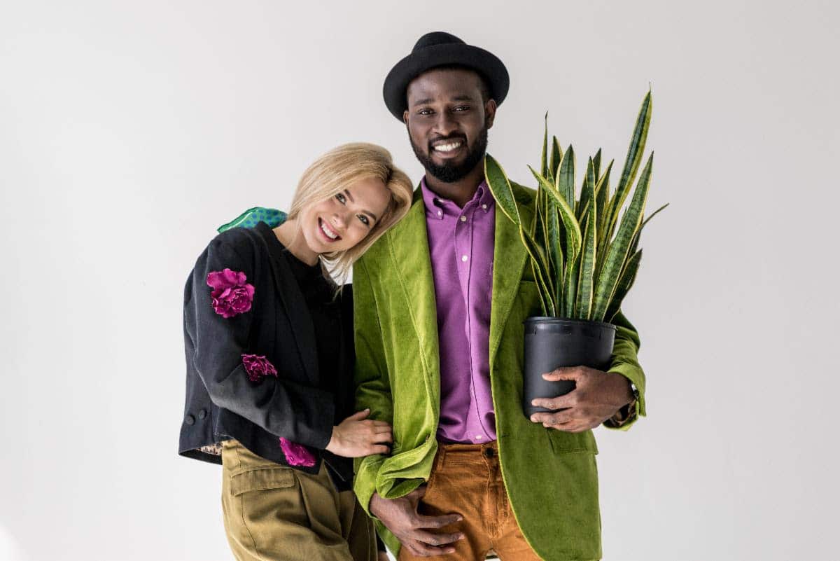 Smiling couple posing together against a plain background, with the man wearing a green jacket and hat while holding a potted snake plant, and the woman leaning on his shoulder.
