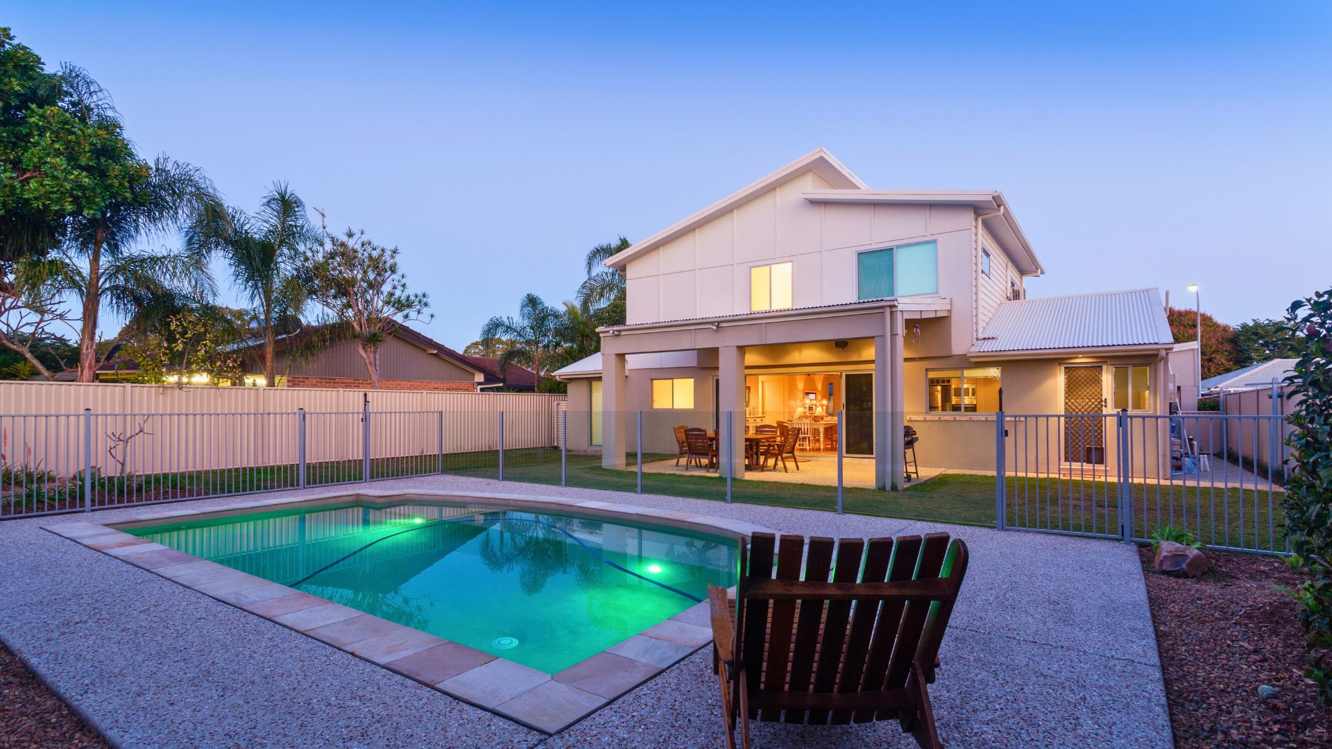 Backyard pool with lighting, pool fence, and covered patio at dusk.
